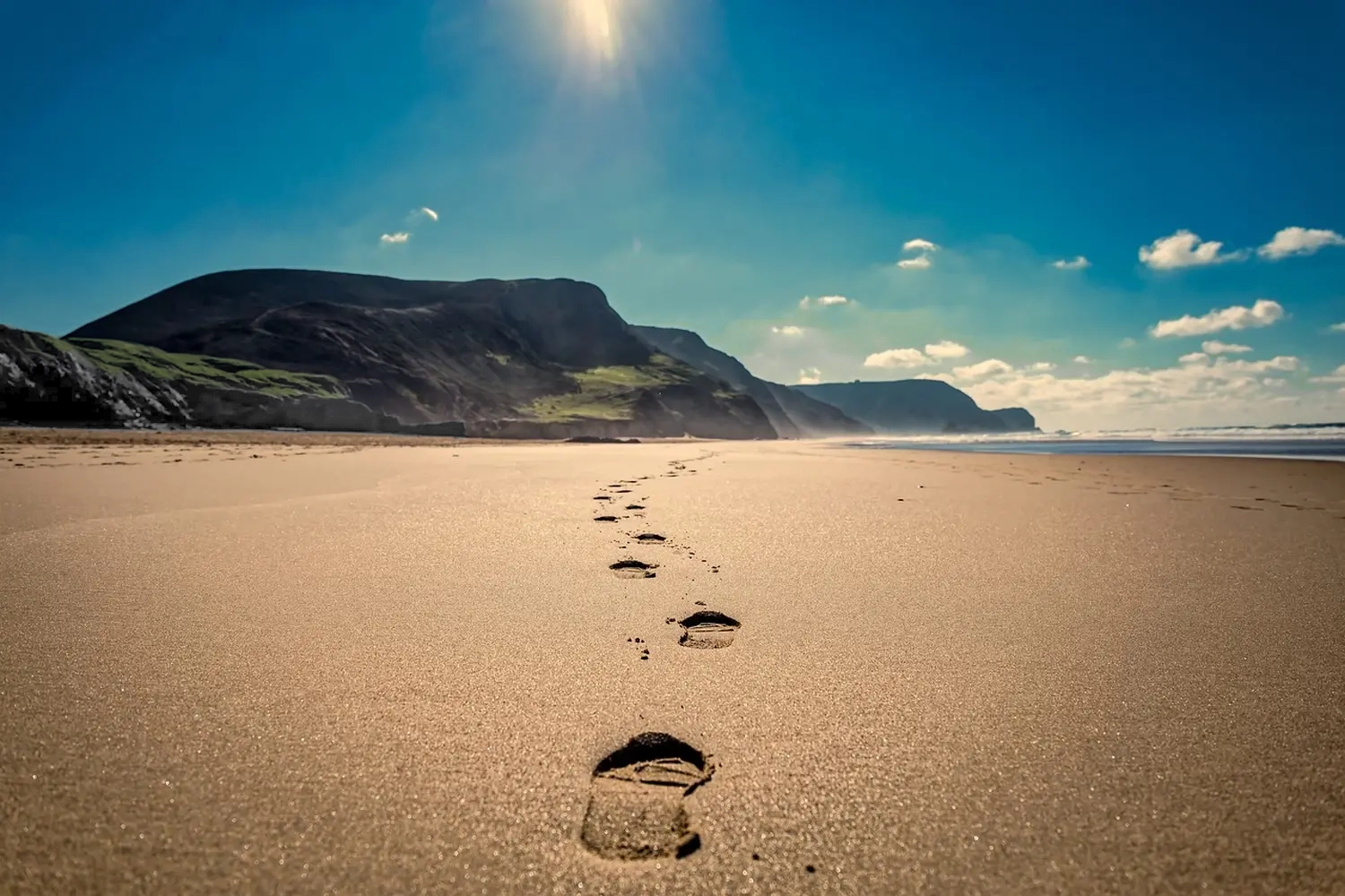 Ein Sandstrand mit deutlichen Fußspuren, die zum Wasser und den Bergen in der Ferne führen, unter einem strahlenden, sonnigen Himmel mit einigen Wolken. Die Sonne scheint hell und lässt den Himmel strahlen. von Hypnose/Mental- & Personaltraining